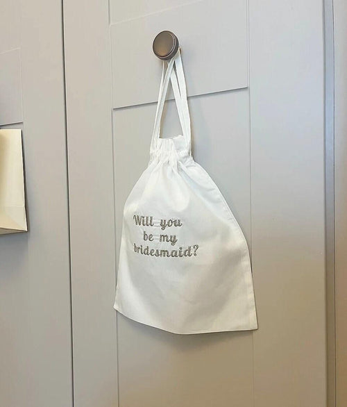White drawstring bag with 'Will you be my bridesmaid?' text hanging on a brass hook.