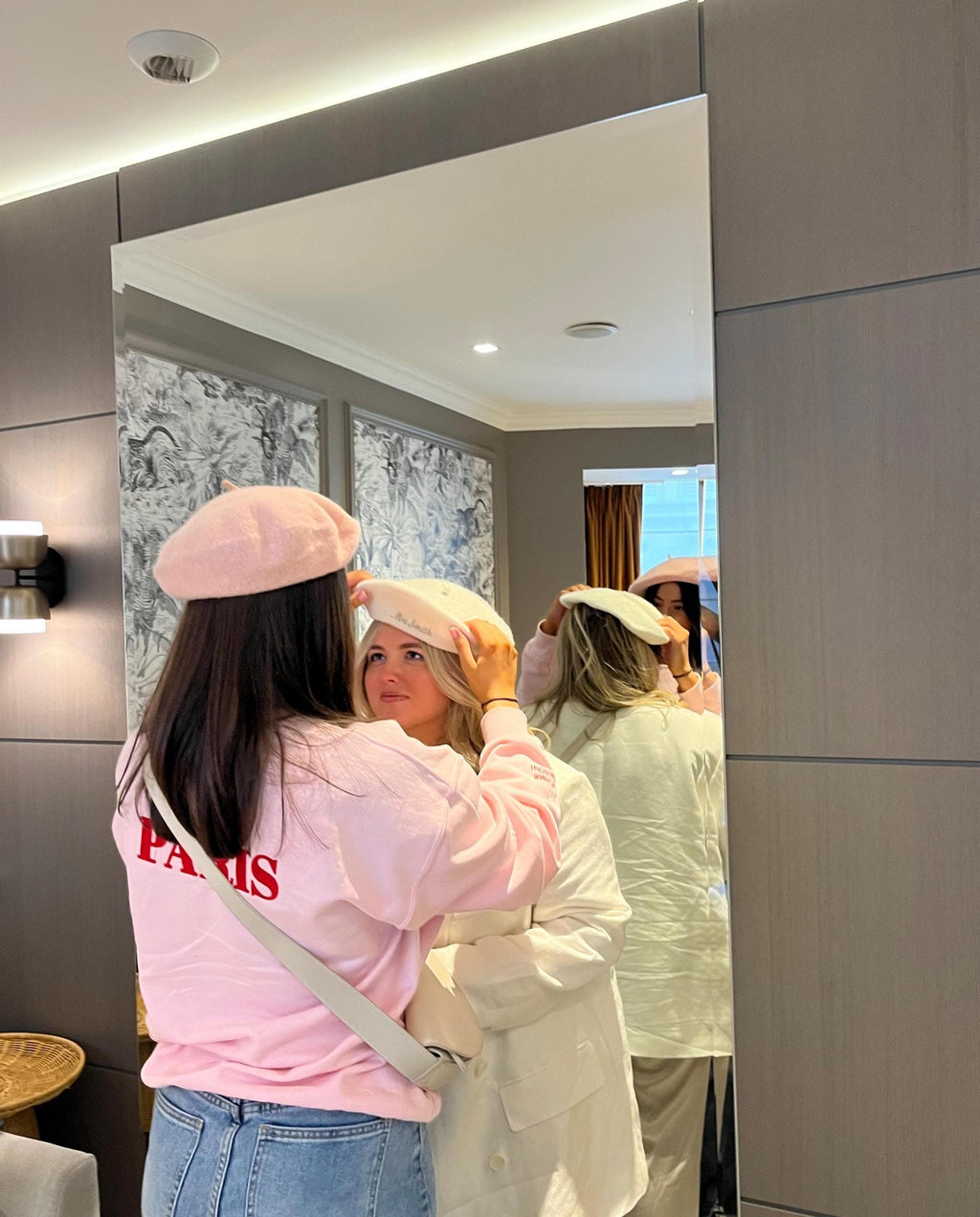 Two women trying on hats in a mirror, with one adjusting the other's hat.
