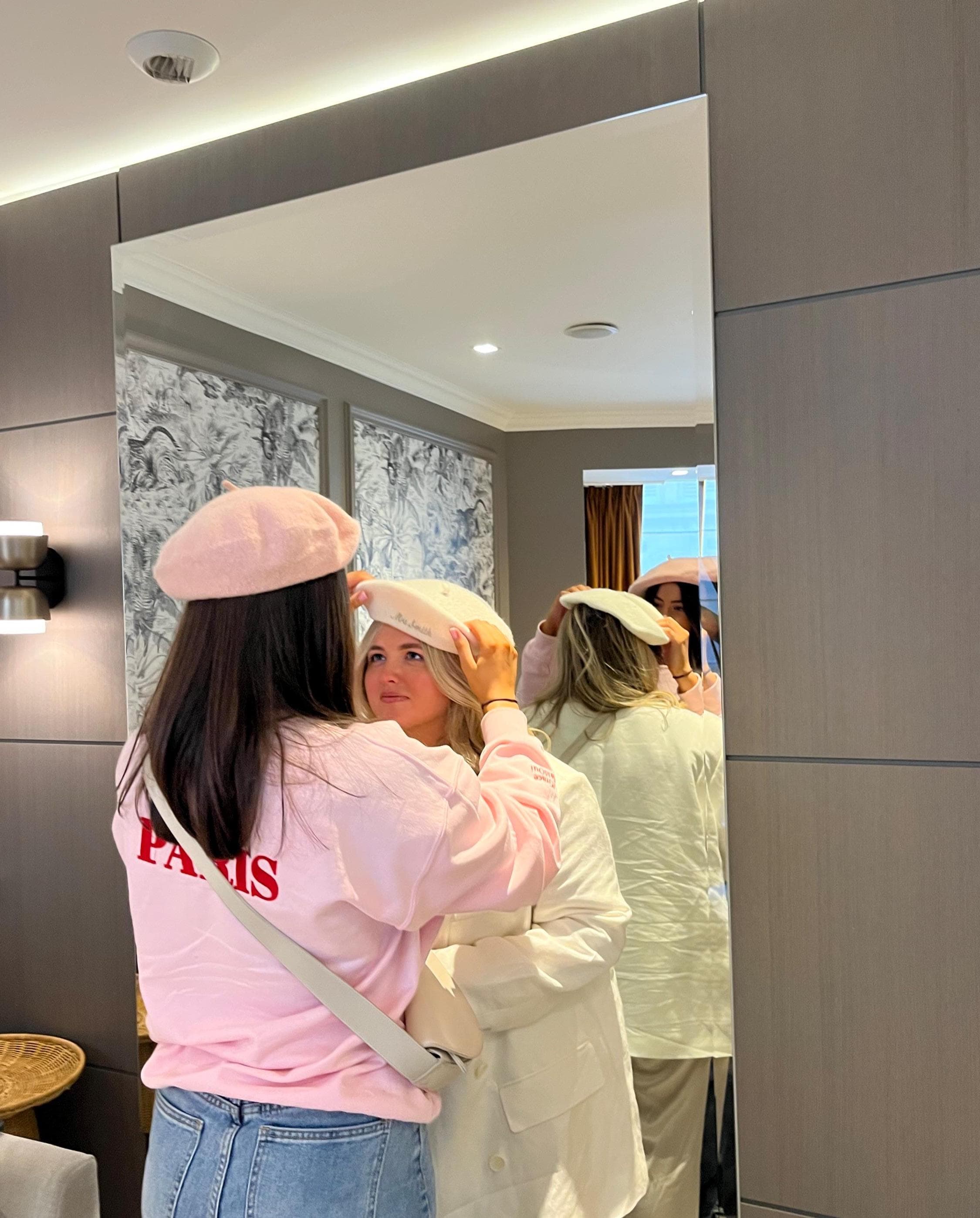 Two women trying on hats in a mirror, with one adjusting the other's hat.
