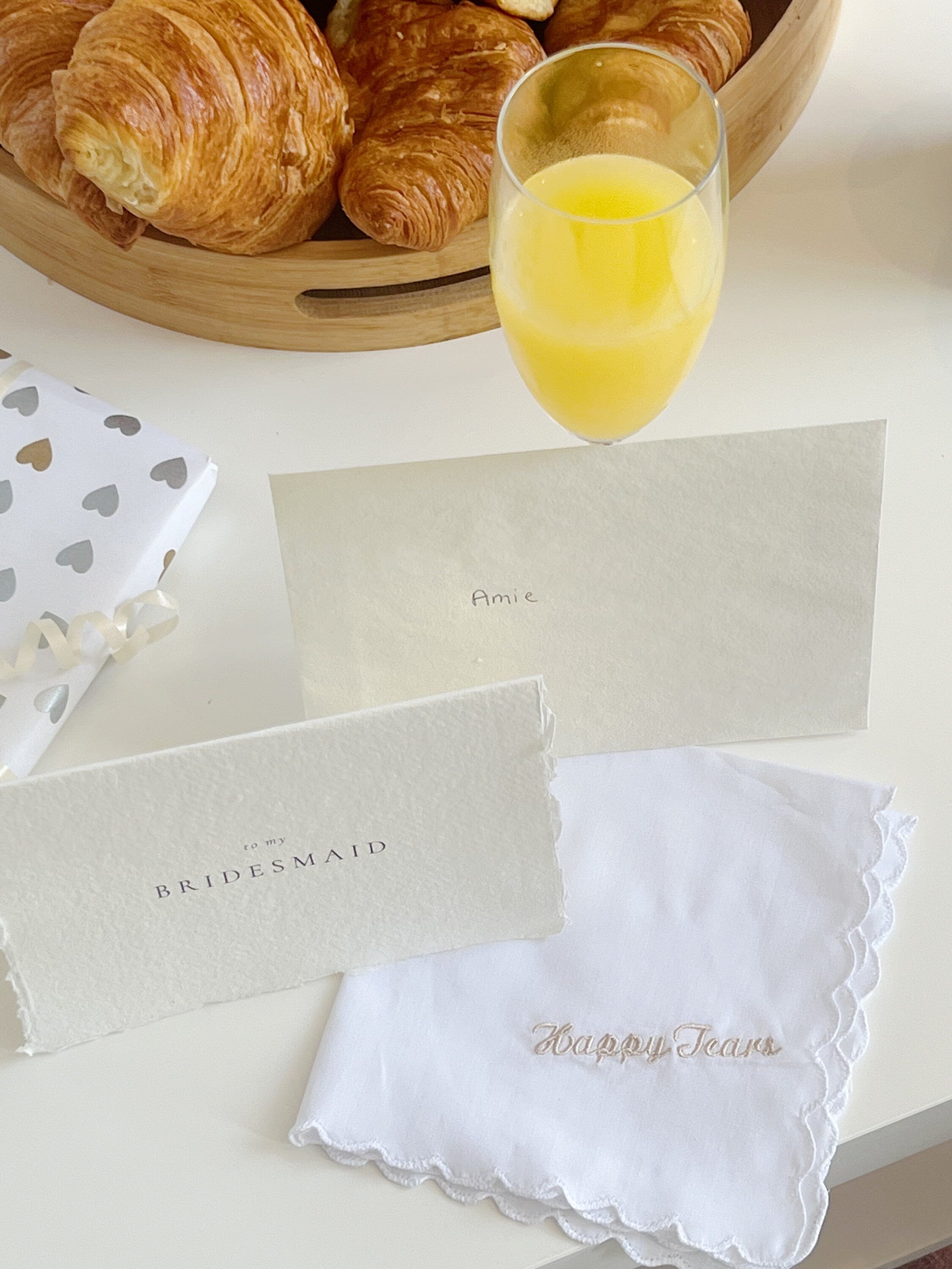 Bouquet of croissants, a glass of orange juice, and a white handkerchief with 'Happy Tears' text on a white surface.