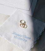 Two gold rings on a white embroidered handkerchief with personalised text.
