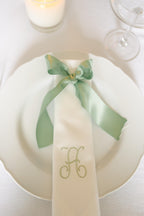 White napkin with green ribbon and monogram on a white plate, set against a white background.